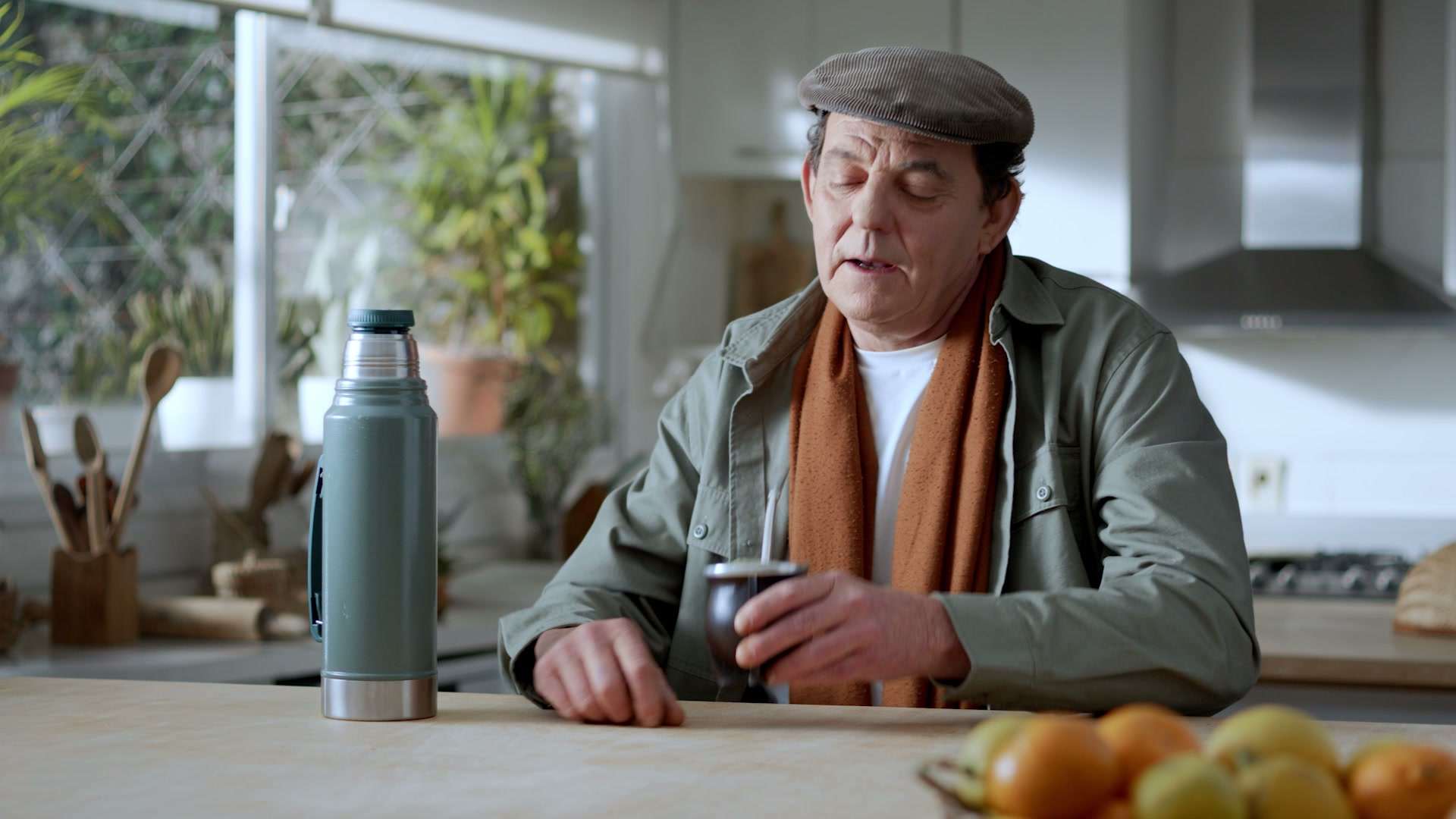Hombre mayor con boina y bufanda naranja, mate en mano, sentado en una cocina
