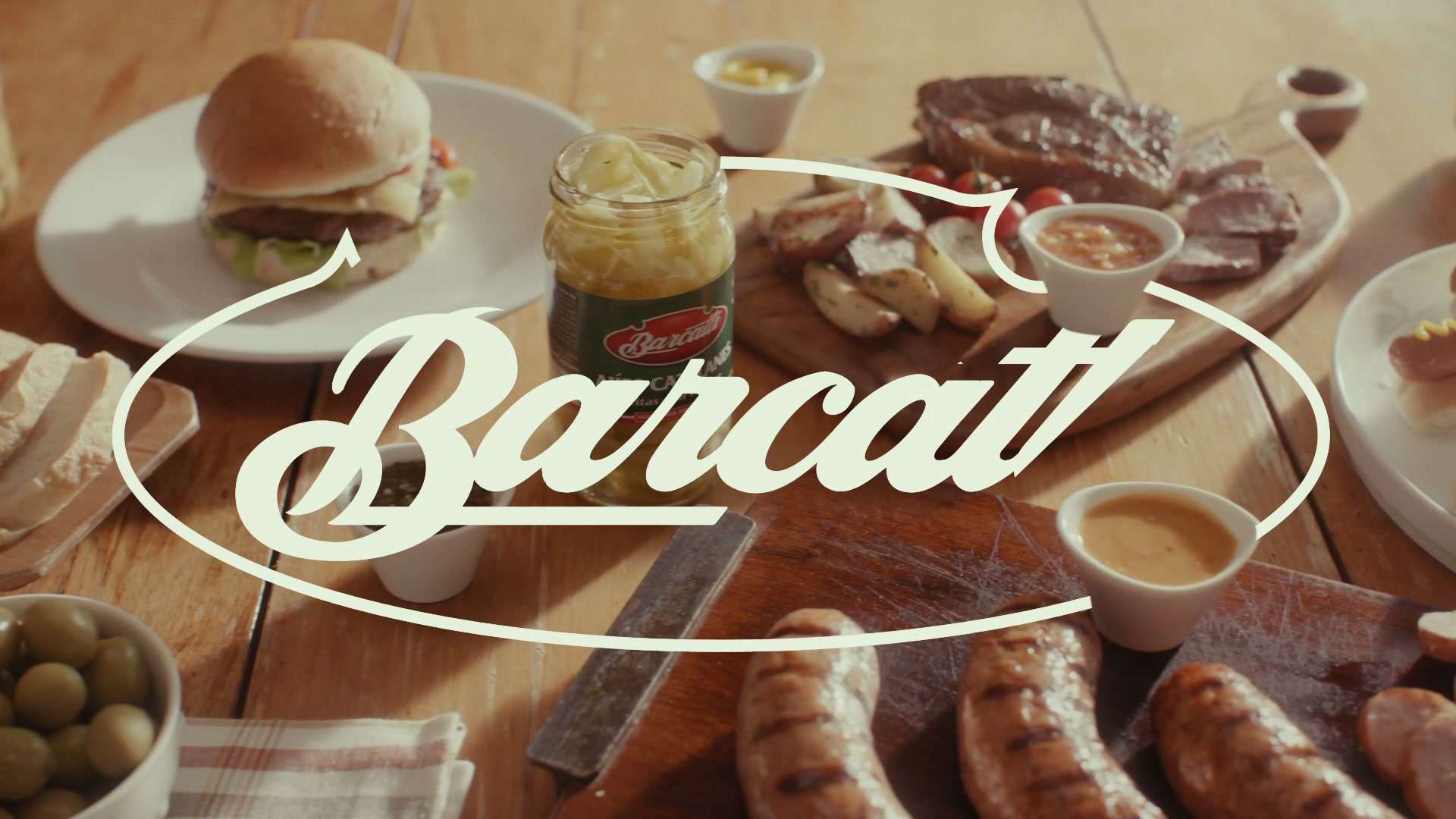 Mesa con hamburguesa, chorizos, aceitunas y salsas, con el logo de Barcatti escrito en el aire
