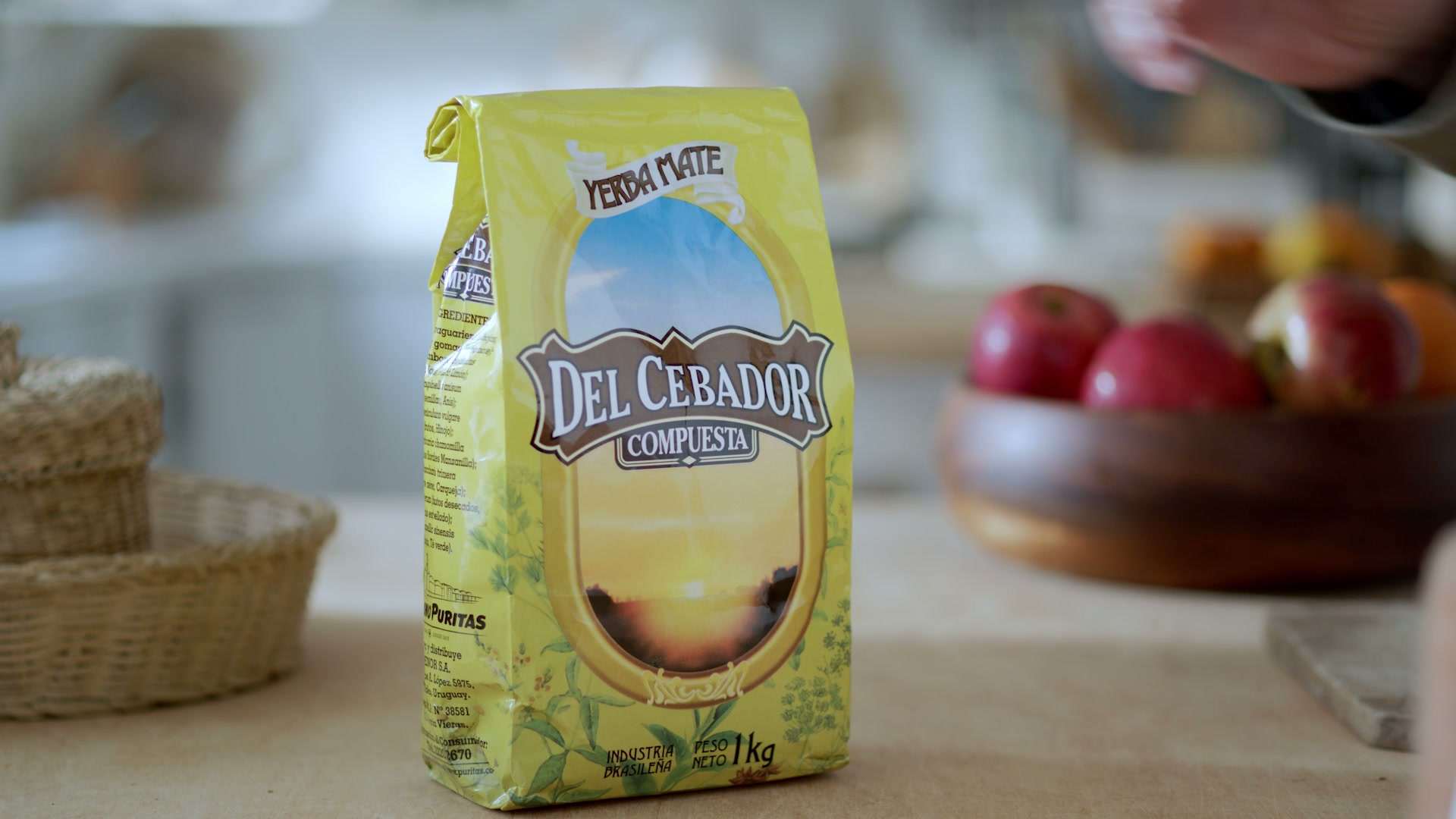 Paquete de yerba Del Cebador Compuesta sobre una mesada, con frutas detrás
